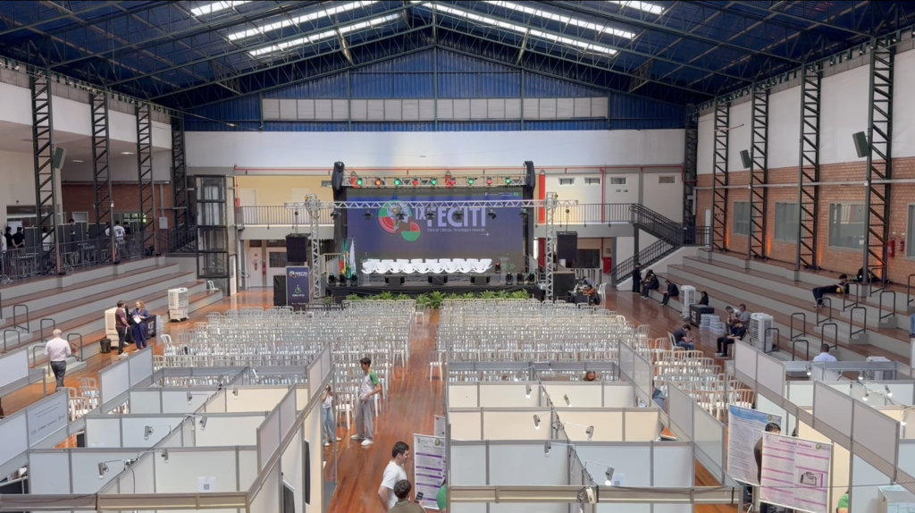 Vista geral do espaço do evento no IFSul em São Leopoldo, com palco, cadeiras e estandes de trabalhos.